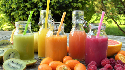 Fünf bunte Smoothies in Glasflaschen mit Strohhalmen stehen auf einem Holztisch im Freien, umgeben von frischem Obst wie Kiwis, Mandarinen, Himbeeren und Grapefruit – symbolisch für die appetitliche, aber oft zuckerreiche Seite verarbeiteter Obstprodukte.